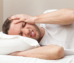 a man wearing a white shirt, laying his head on a white pillow, and holding the left side of his head in chronic pain