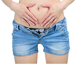 a woman wearing unzipped denim shorts with her two hands touching her abdomen in pain